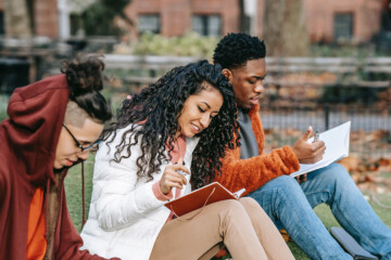 Three college students sitting outside on a college campus talking and taking ntoes.