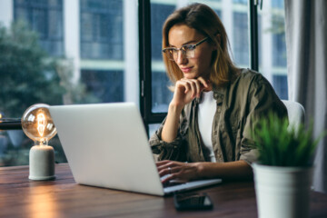 A woman learning about customer journey mapping with shoulder-length hair wearing glasses sits at a desk looking at a laptop with a large window behind her.