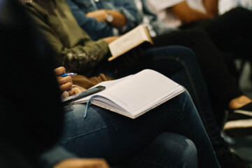 A women sitting at a conference while taking notes with a notebook, pen and her phone.