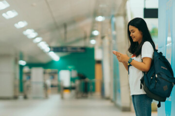 An Asian college student with a blue backpack stands in a school hallway while looking at her cell phone.