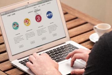 A man sits at a wood table with a cup of espresso and types on his laptop.