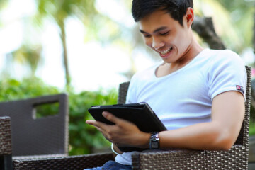 A young man in a white t-shirt sits at a patio table drinking iced tea while scrolling through an iPad.