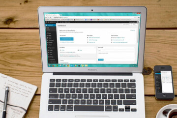 Laptop on a wood desk with pad of paper, pen, smartphone, and cup of coffee.