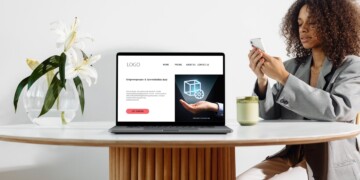 A woman with curly brown hear wearing a grey jacket looks at her smartphone while she sits at a white table with a laptop open to a landing page design.