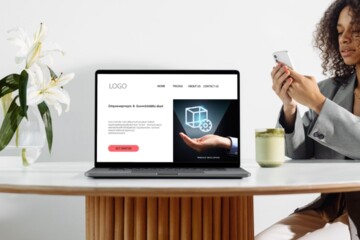 A woman with curly brown hear wearing a grey jacket looks at her smartphone while she sits at a white table with a laptop open to a landing page design.