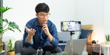 An Asian man in a blue dress shirt sits in his living room an creates a podcast with his smartphone.