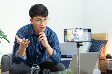 An Asian man in a blue dress shirt sits in his living room an creates a podcast with his smartphone.