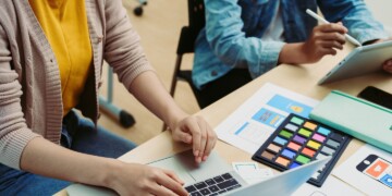 Two female coworkers working at a shared desk with laptops.