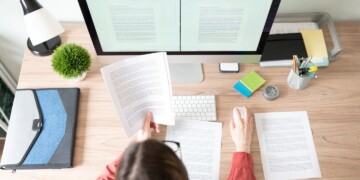 Person sitting at a desk, reading a print out and working on their desktop.